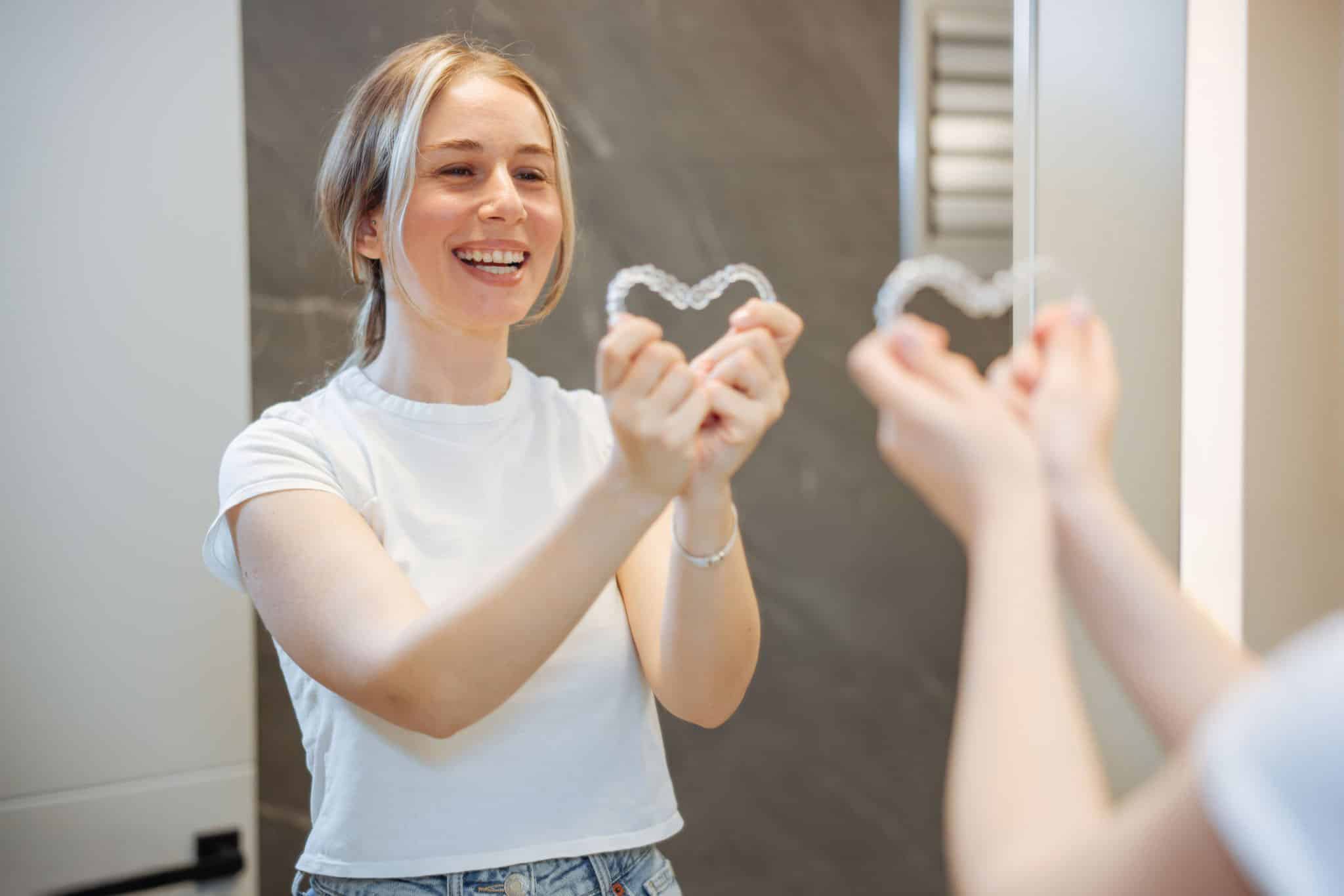 Alt text<br />
Smiling woman holding clear aligners in heart shape, showing love for her orthodontic treatment in bathroom mirror<br />