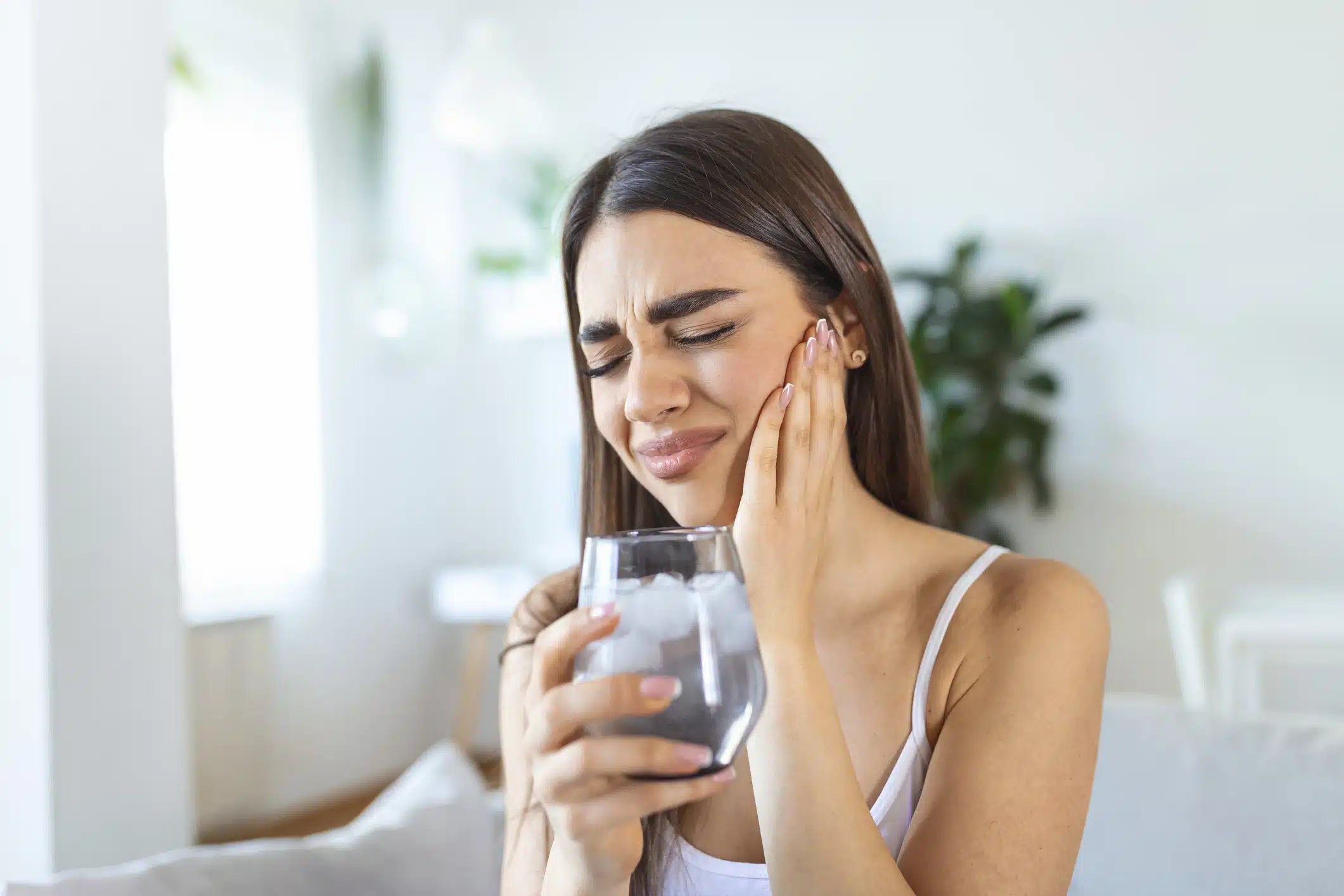 Young woman with sensitive teeth and hand holding glass of cold water with ice.