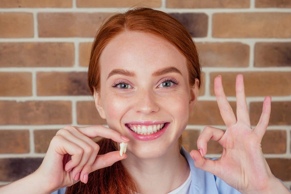 a woman smiling and holding her wisdom tooth that was recently extracted