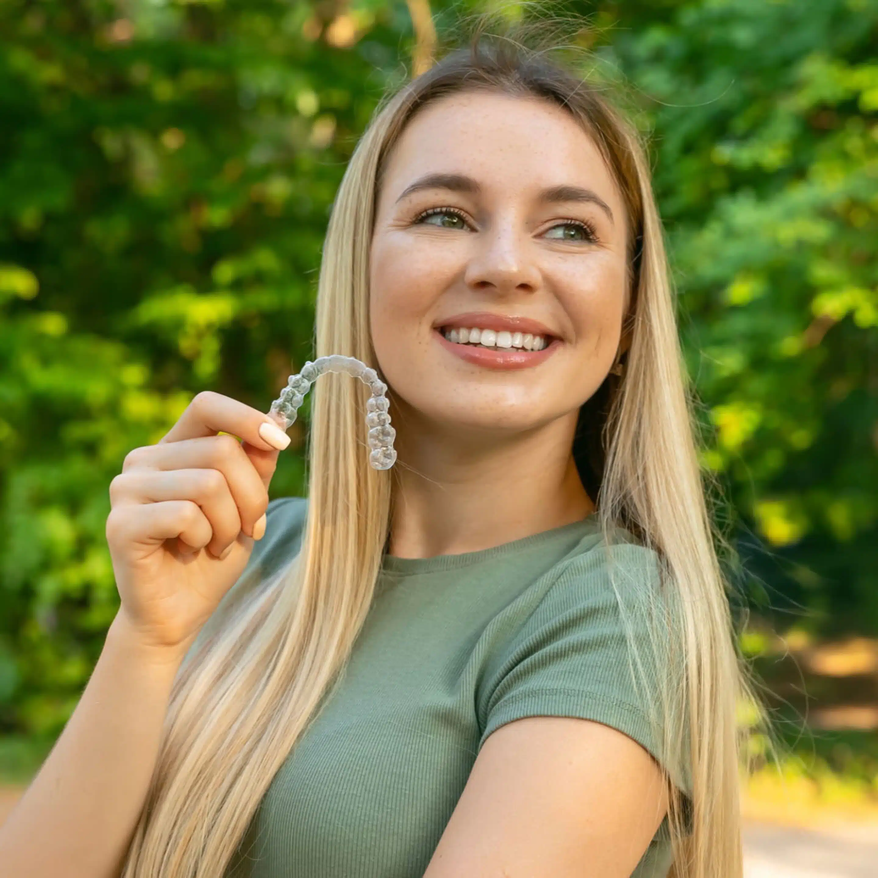 Smiling woman holding a clear aligner outdoors, showing straight teeth after orthodontic treatment<br />