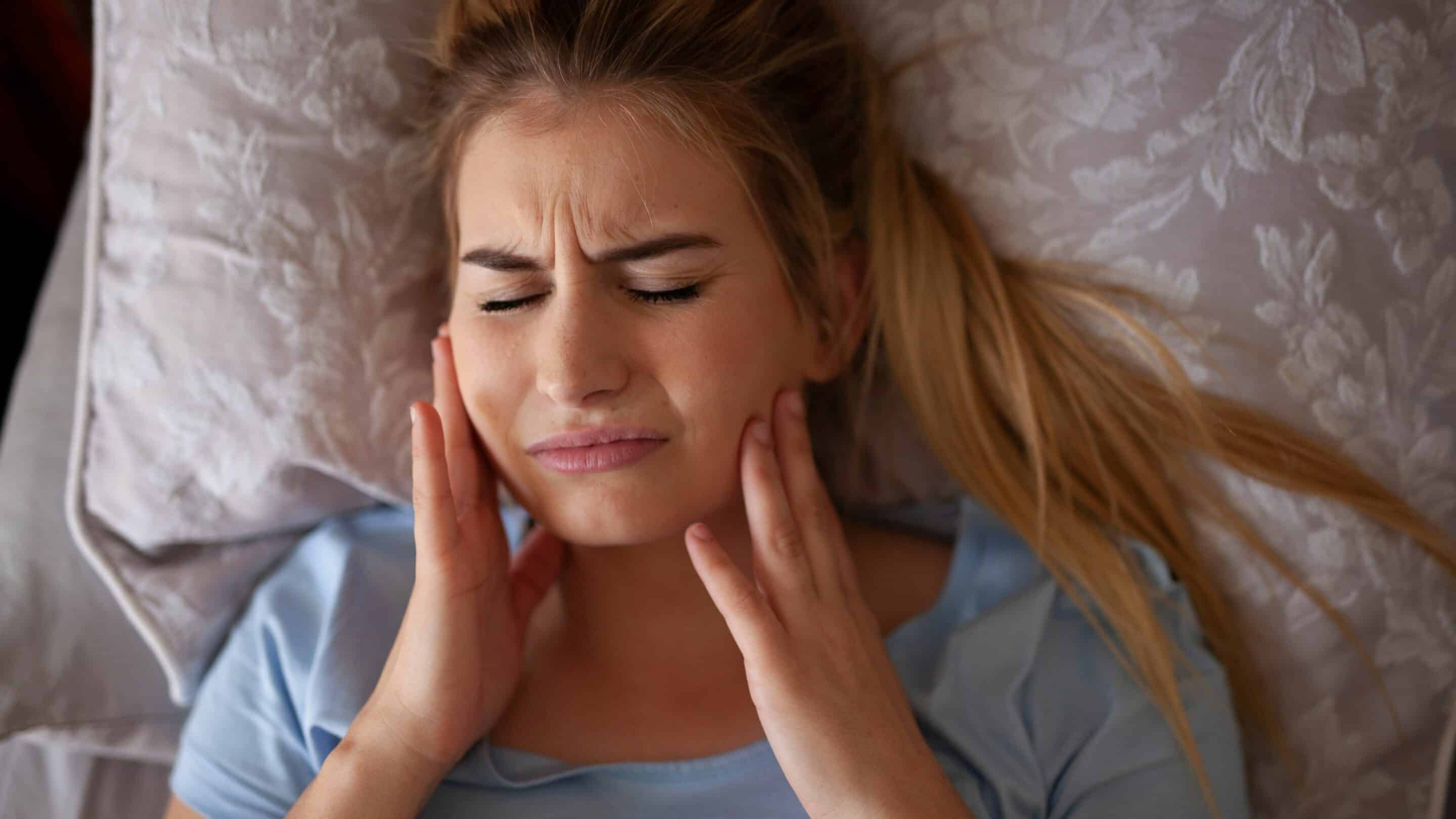 Woman lying in bed holding jaw showing signs of TMJ pain.